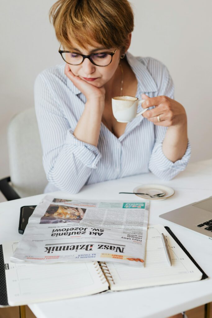 A thoughtful woman reads a newspaper while enjoying coffee at an indoor workspace.